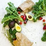 Avocado, kale, and more fresh produce artfully arranged on a marble surface.